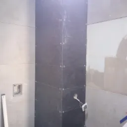 Wnętrze łazienki w trakcie prac wykończeniowych: pionowa kolumna obłożona ciemnymi płytkami z krzyżykami dystansowymi, jasne płytki na ścianach, widoczne otwory pod instalacje, poziomica oparta...