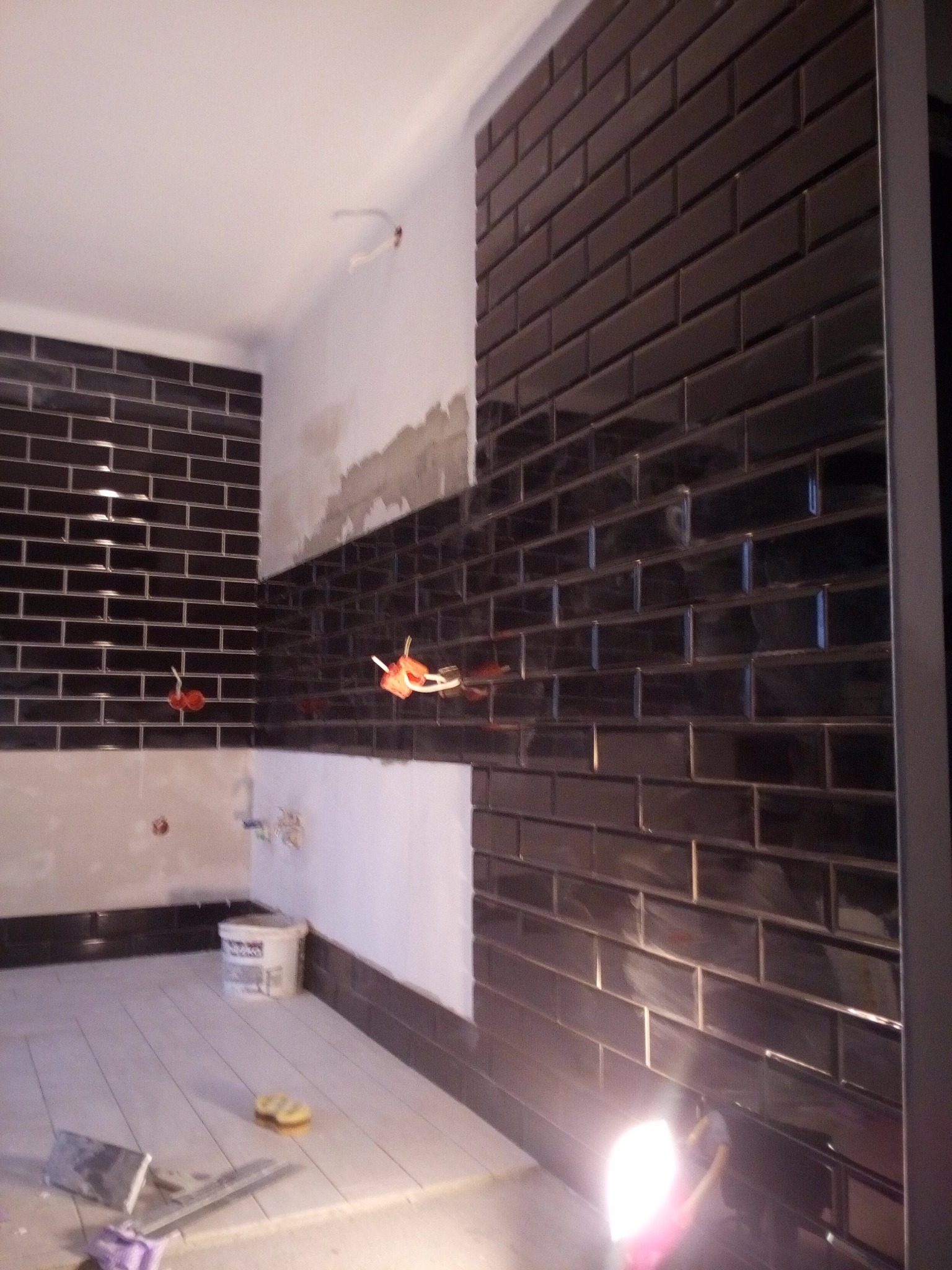 Częściowo wykończona łazienka z czarnymi, błyszczącymi płytkami w stylu cegiełek na ścianach, widoczne instalacje elektryczne, wiadro z zaprawą oraz narzędzia na podłodze z jasnych płytek.