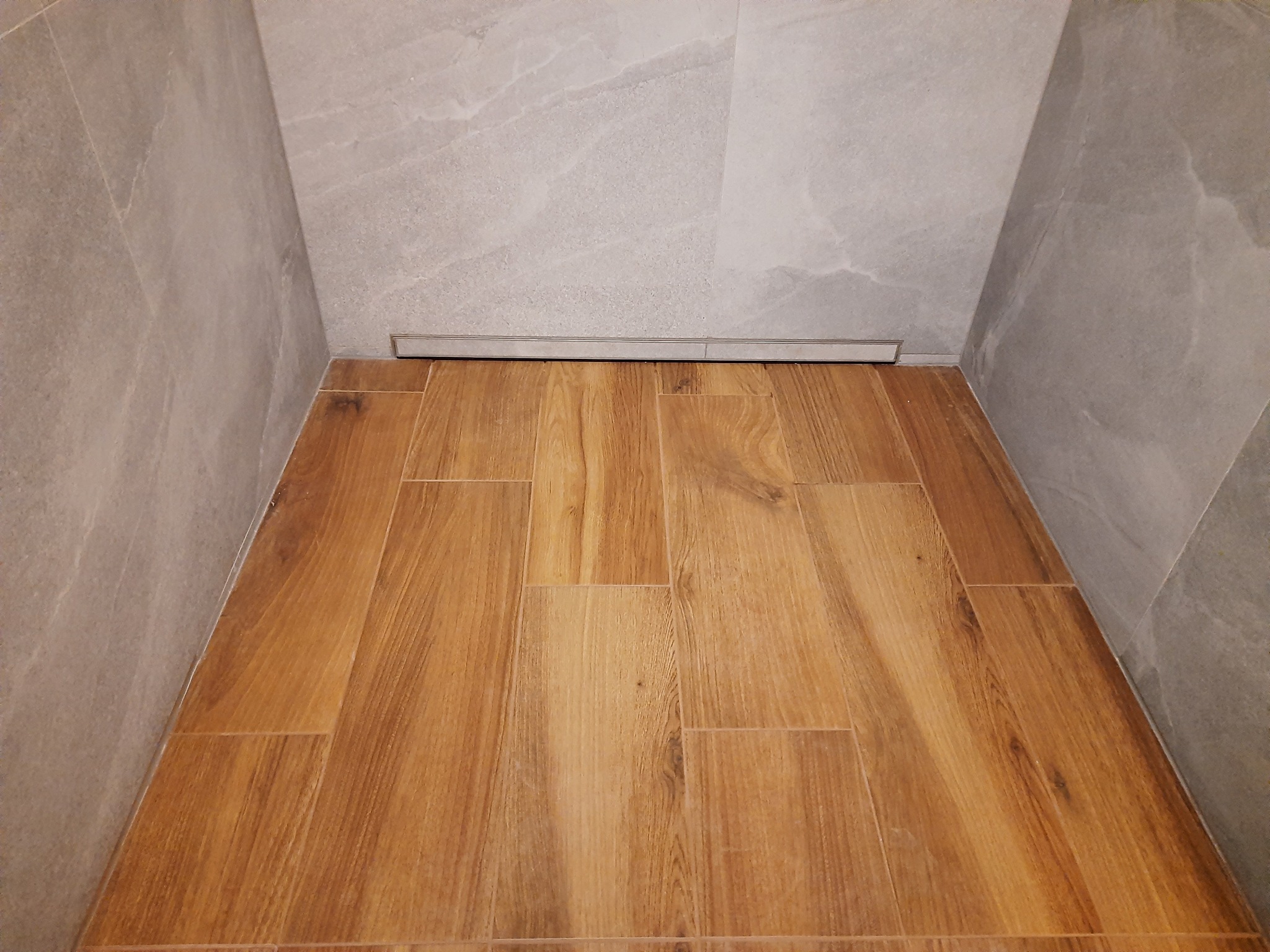 Wykończona kabina prysznicowa z drewnopodobnymi płytkami podłogowymi i szarymi płytkami ściennymi, widoczny odpływ liniowy.