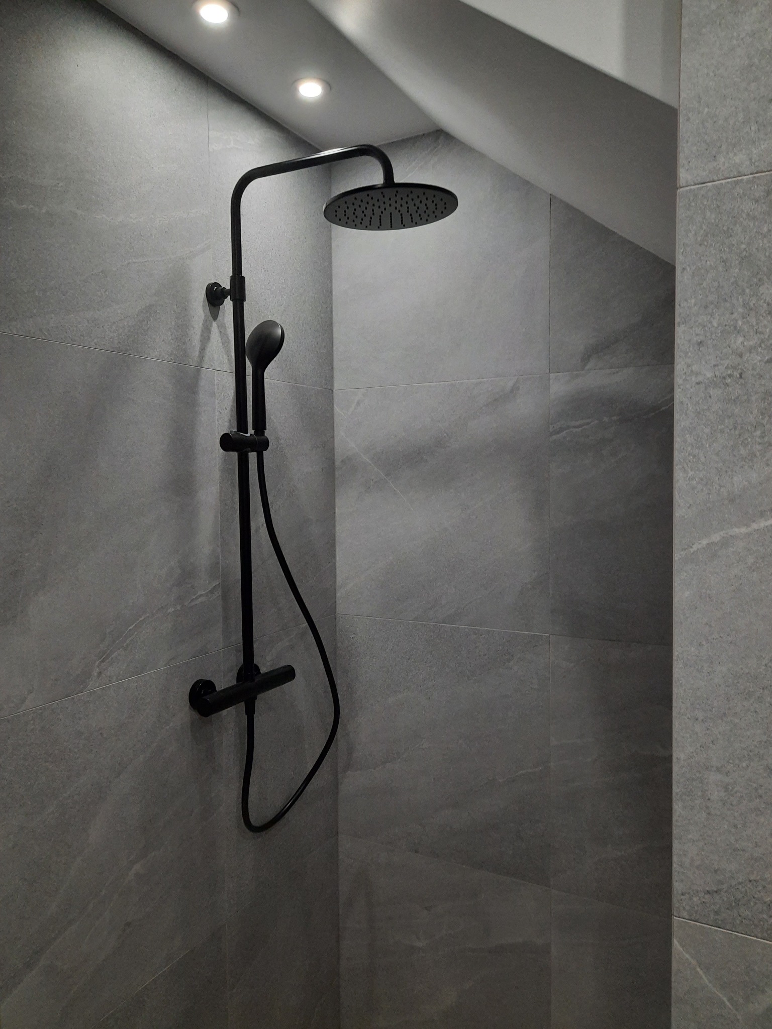 Nowoczesna kabina prysznicowa z czarną armaturą i szarymi płytkami imitującymi kamień, oświetlona punktowymi lampami sufitowymi.