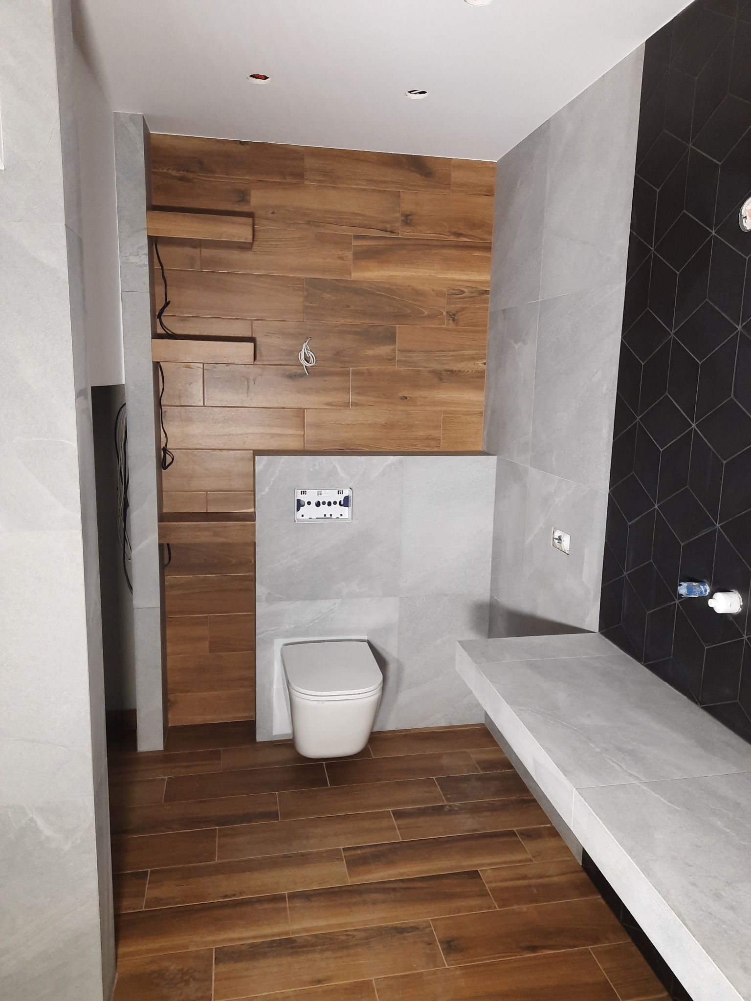Wnętrze łazienki z drewnopodobnymi i szarymi płytkami, wisząca toaleta i zabudowa ze stelażem, częściowo wykończona ściana z przewodami.