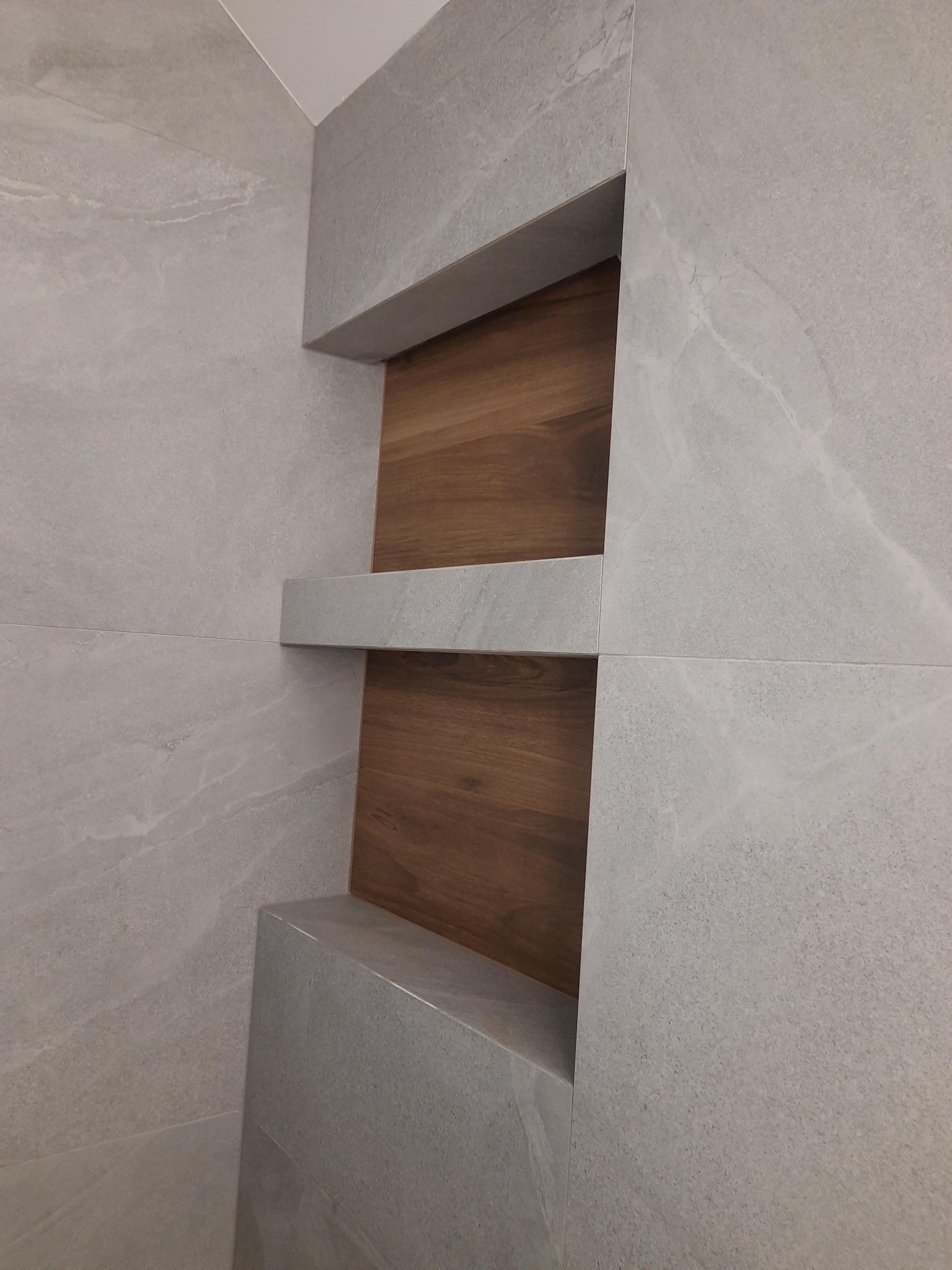 Wnęka z dwiema półkami wyłożona szarymi płytkami imitującymi kamień z drewnianym tłem.