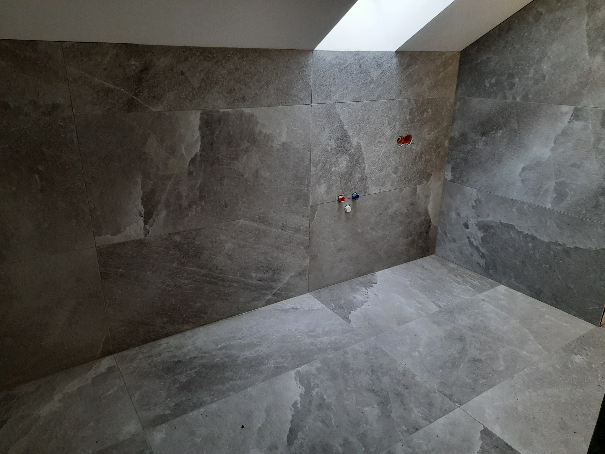 Wnętrze łazienki w trakcie prac wykończeniowych, widoczne szare płytki imitujące marmur na ścianach i podłodze, otwory na instalacje wodne i elektryczne, oraz okno dachowe.