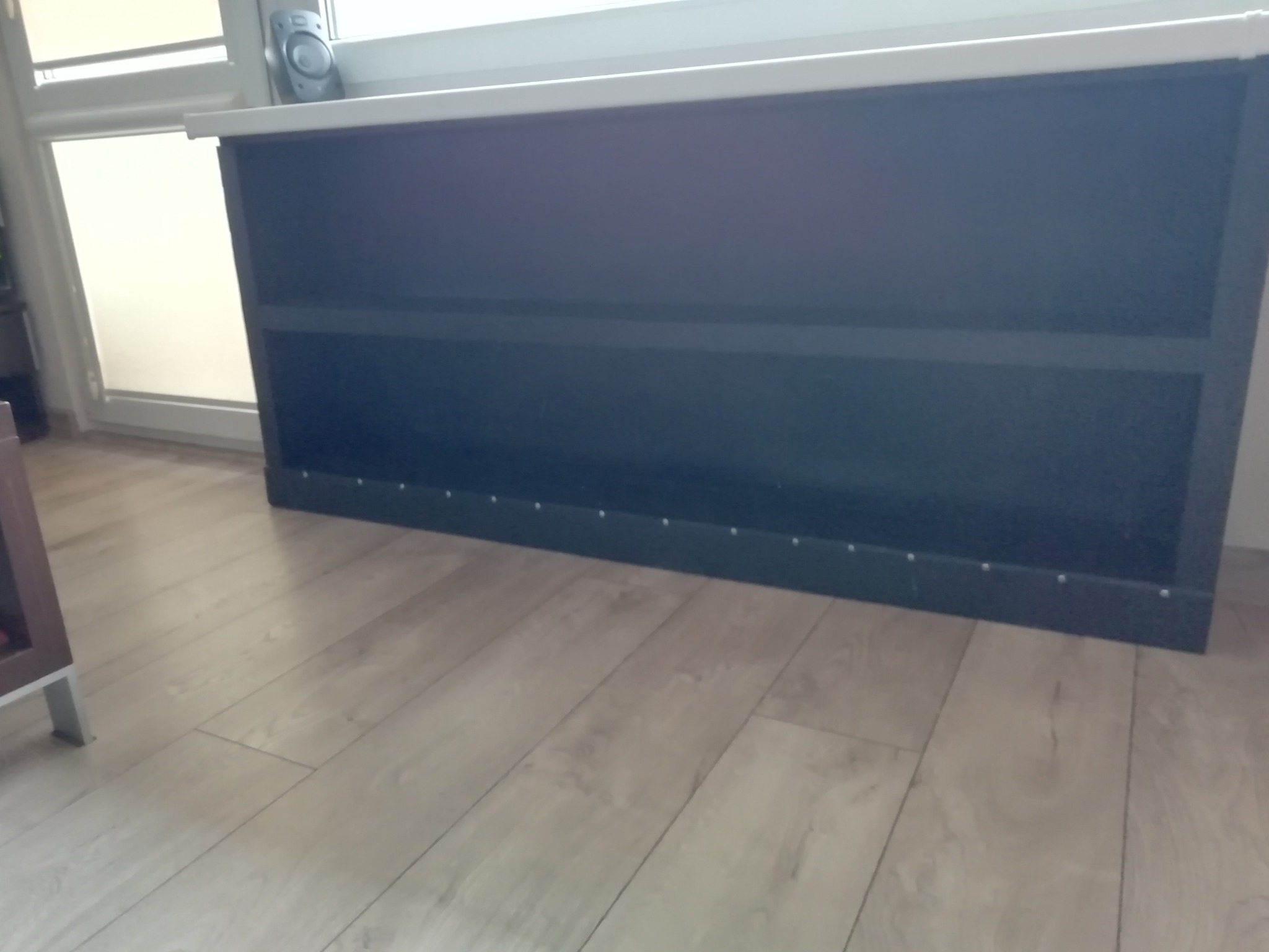 Czarna zabudowa grzejnika z drewnianymi listwami pod parapetem na tle okna.