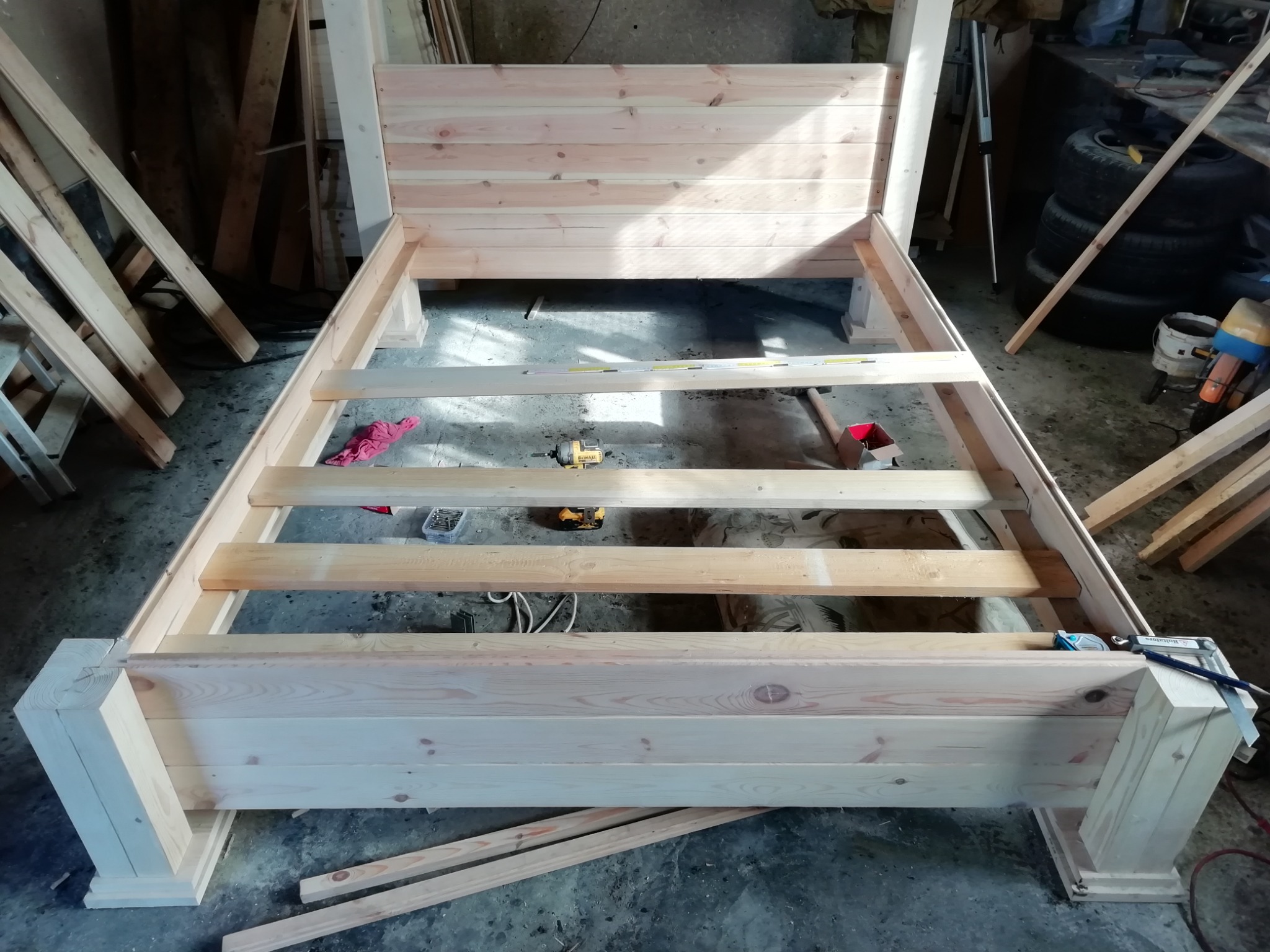 Rama łóżka z surowego drewna w trakcie montażu, widoczne narzędzia i elementy konstrukcyjne, warsztatowe otoczenie.