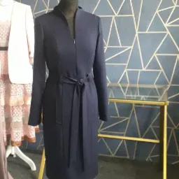 Elegancka, granatowa sukienka z długim rękawem, z paskiem wiązanym w talii, zapinana na zamek, prezentowana na manekinie na tle ściany z geometrycznym wzorem w odcieniach szarości i złota. Obok...