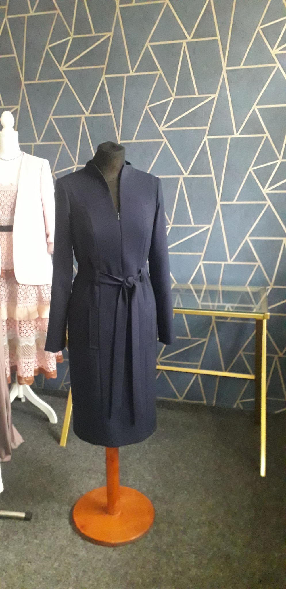 Elegancka, granatowa sukienka z długim rękawem, z paskiem wiązanym w talii, zapinana na zamek, prezentowana na manekinie na tle ściany z geometrycznym wzorem w odcieniach szarości i złota. Obok...