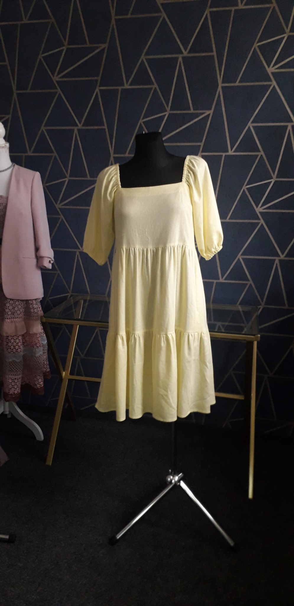 Jasnożółta, bawełniana sukienka z kwadratowym dekoltem i krótkimi, bufkowatymi rękawami, zaprezentowana na czarnym manekinie na tle granatowej ściany z geometrycznym wzorem; obok widać fragment...