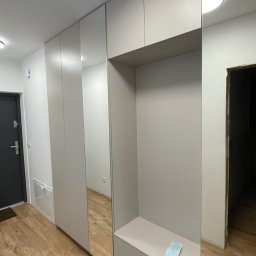 Roman Jaśkiewicz JASK-MEBLE - Jasna zabudowa przedpokoju z szafami, siedziskiem i lustrami, widok z perspektywy podłogi, na siedzisku leży kartka.