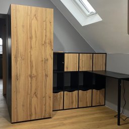 Roman Jaśkiewicz JASK-MEBLE - Zabudowa pod skosem na poddaszu: szafa z frontami imitującymi drewno i regał zamykany na czarno z drewnianymi frontami, połączony z czarnym biurkiem.