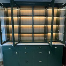 Roman Jaśkiewicz JASK-MEBLE - Zielony kredens z przeszklonymi drzwiczkami i wewnętrznym oświetleniem LED, złote uchwyty, płytki podłogowe w romby.