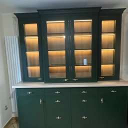 Roman Jaśkiewicz JASK-MEBLE - Zielony, lakierowany kredens z przeszkloną nadstawką, podświetlany od wewnątrz, z delikatnymi złotymi uchwytami, ustawiony na beżowej podłodze z płytek.