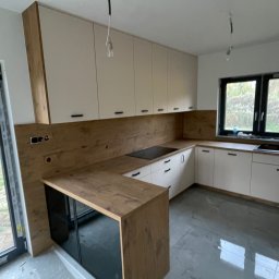 Roman Jaśkiewicz JASK-MEBLE - Nowoczesna kuchnia z białymi szafkami i drewnianym blatem, widoczne kable wystające z sufitu, sugerujące niedokończony montaż oświetlenia, podłoga w trakcie wykończenia.