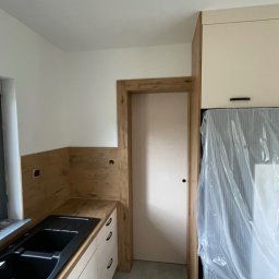 Roman Jaśkiewicz JASK-MEBLE - Nowoczesna kuchnia z białymi szafkami, czarnym zlewozmywakiem i blatem imitującym drewno, z widocznym fragmentem lodówki zabezpieczonej folią ochronną i drzwiami w drewnianej ościeżnicy.