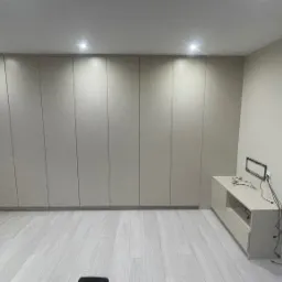 Beżowa szafa wnękowa z przesuwnymi drzwiami, zajmująca całą ścianę, oświetlona punktowymi lampami sufitowymi, na podłodze jasne panele, obok szafki RTV z czarną ramką na ścianie i widocznymi kablami.