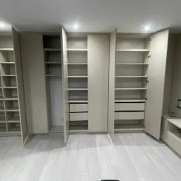 Jasna, nowoczesna garderoba z otwartymi półkami i szufladami, z widocznymi zawiasami i szczelinami między drzwiami, na tle jasnej podłogi i ściany.