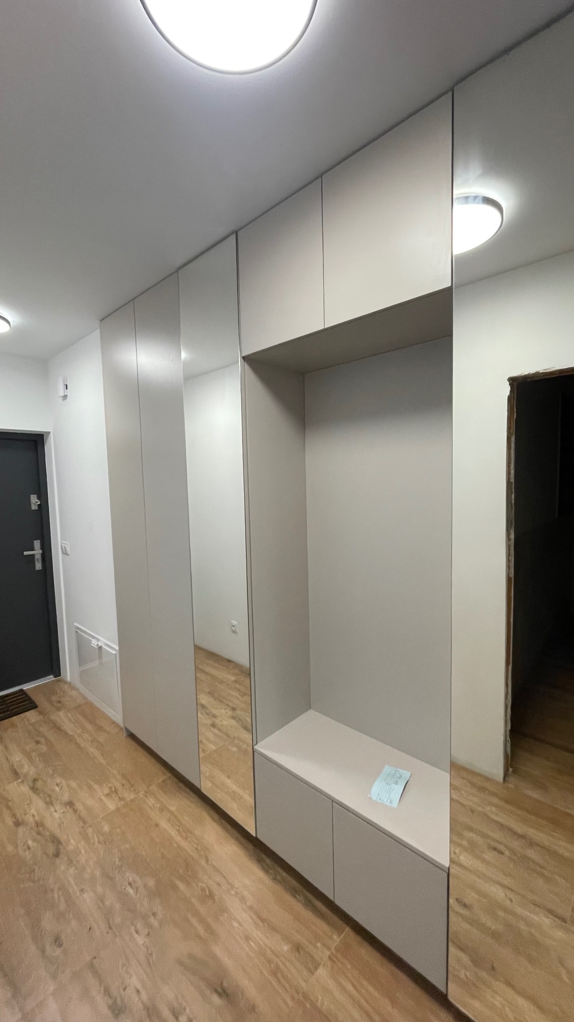 Jasna zabudowa przedpokoju z szafami, siedziskiem i lustrami, widok z perspektywy podłogi, na siedzisku leży kartka.