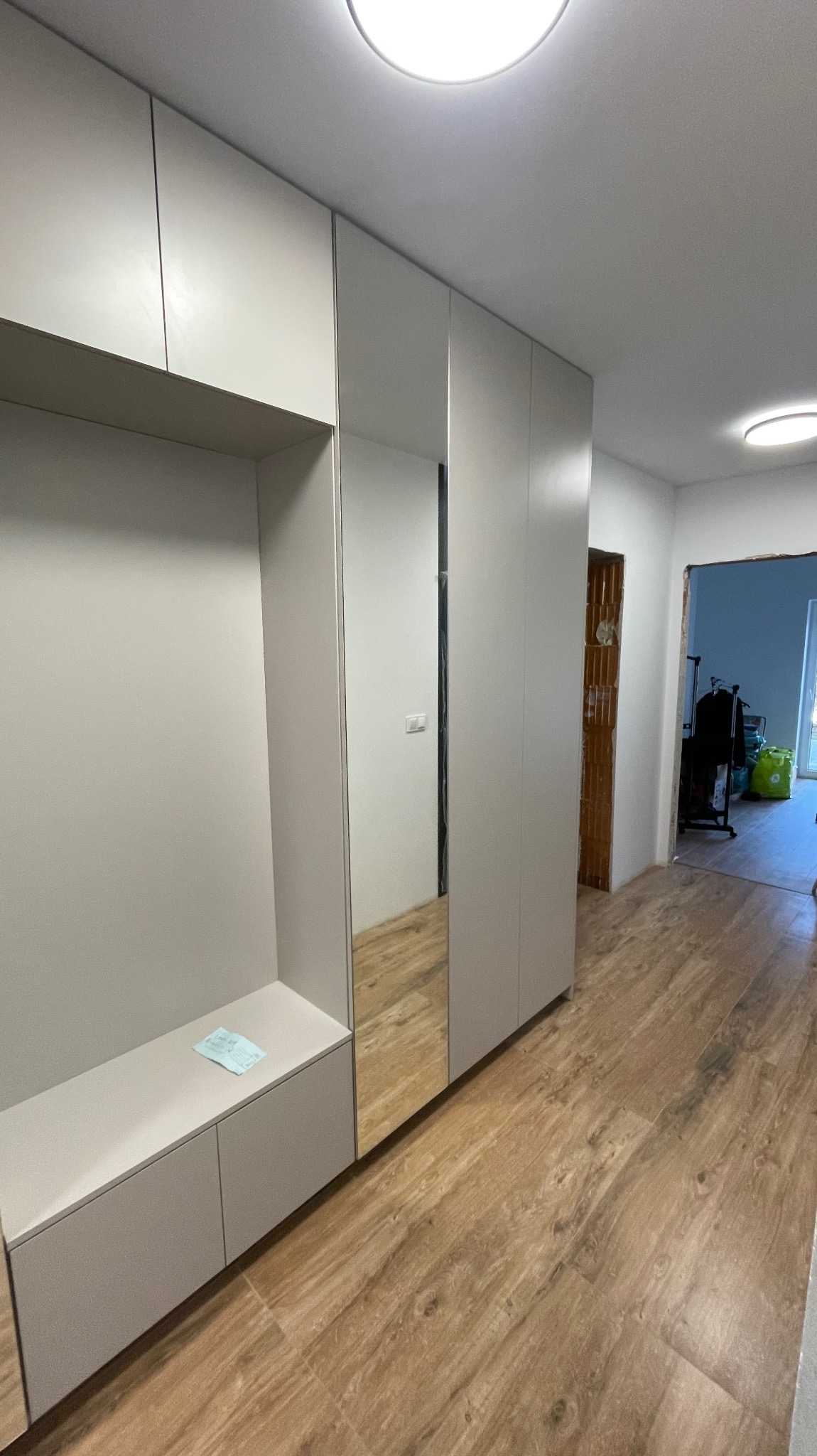 Jasnoszara szafa wnękowa z lustrem na korytarzu, widoczny fragment podłogi imitującej drewno i dokument leżący na półce.