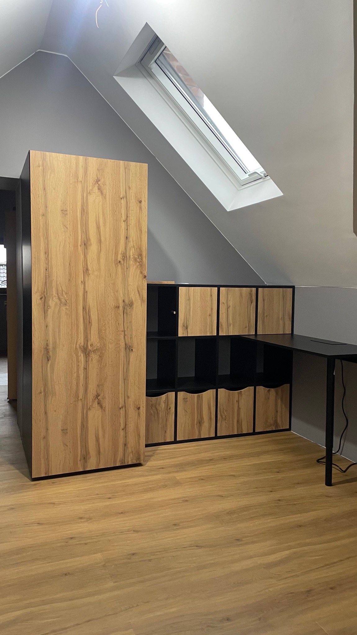 Zabudowa pod skosem na poddaszu: szafa z frontami imitującymi drewno i regał zamykany na czarno z drewnianymi frontami, połączony z czarnym biurkiem.