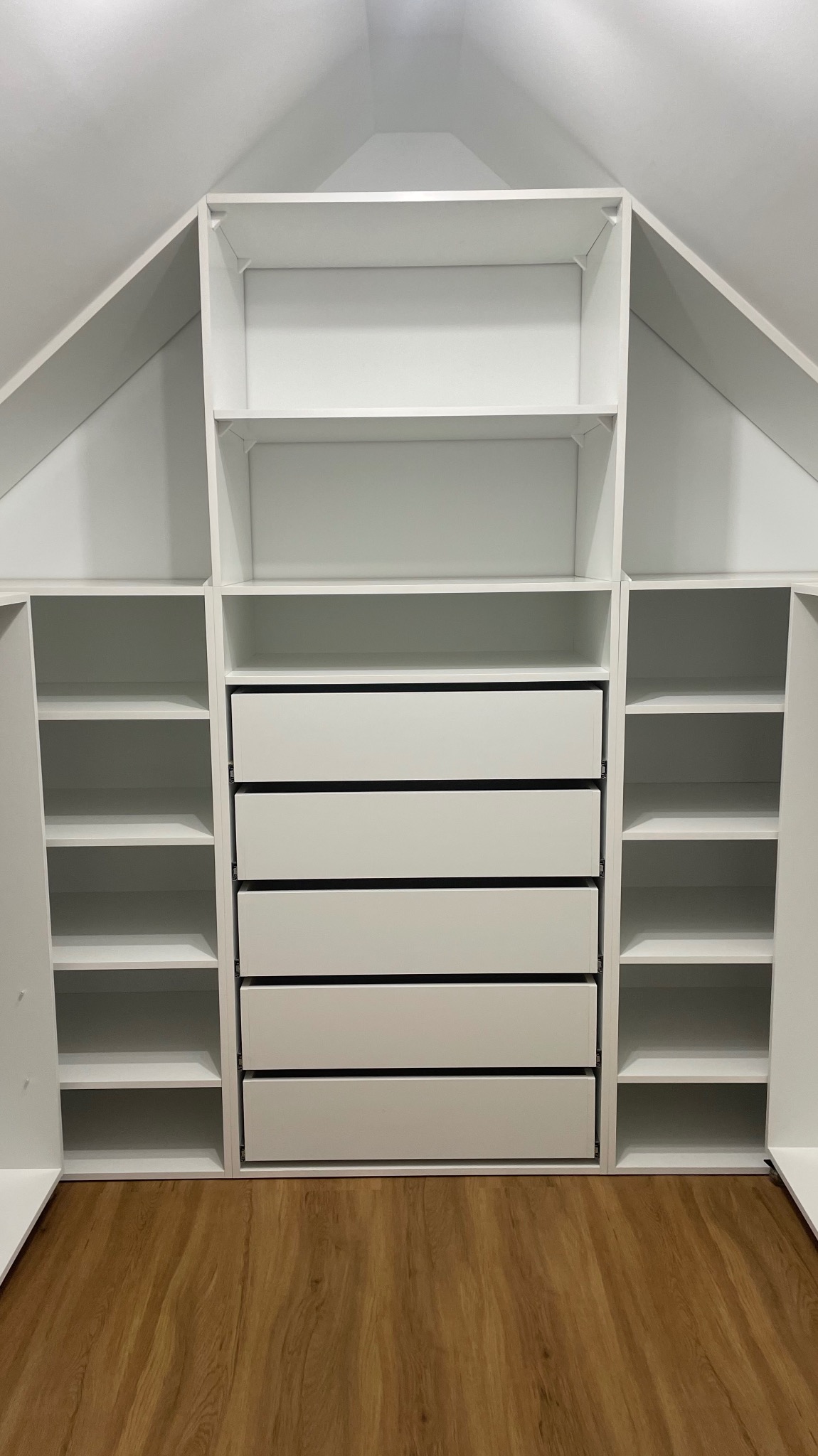 Biała szafa wnękowa z półkami i szufladami, dopasowana do skosu poddasza, widoczna na tle drewnianej podłogi.