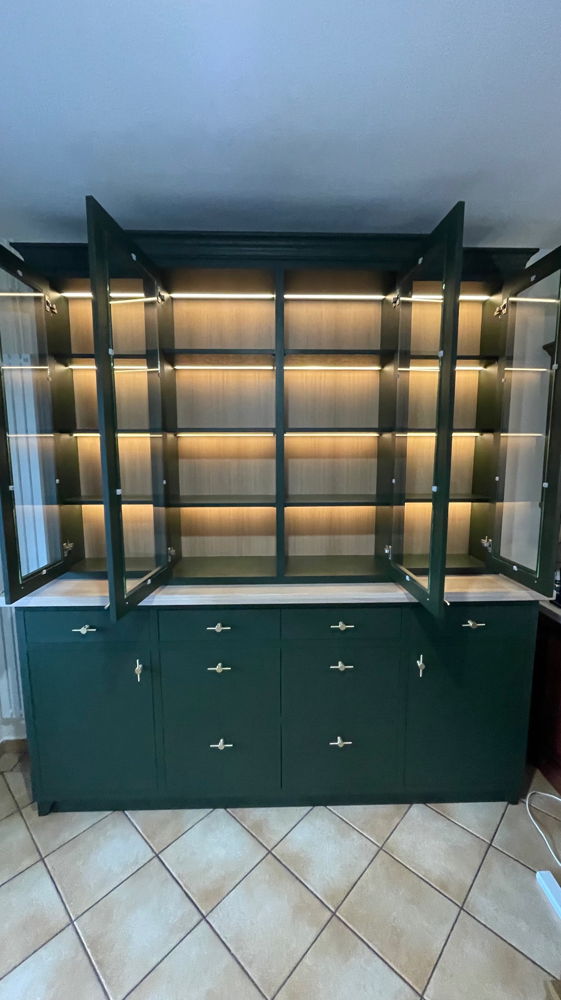 Zielony kredens z przeszklonymi drzwiczkami i wewnętrznym oświetleniem LED, złote uchwyty, płytki podłogowe w romby.