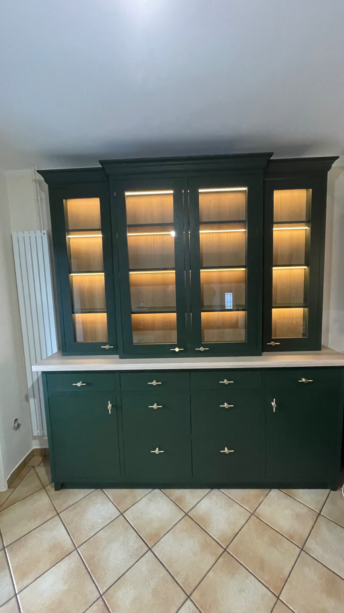 Zielony, lakierowany kredens z przeszkloną nadstawką, podświetlany od wewnątrz, z delikatnymi złotymi uchwytami, ustawiony na beżowej podłodze z płytek.