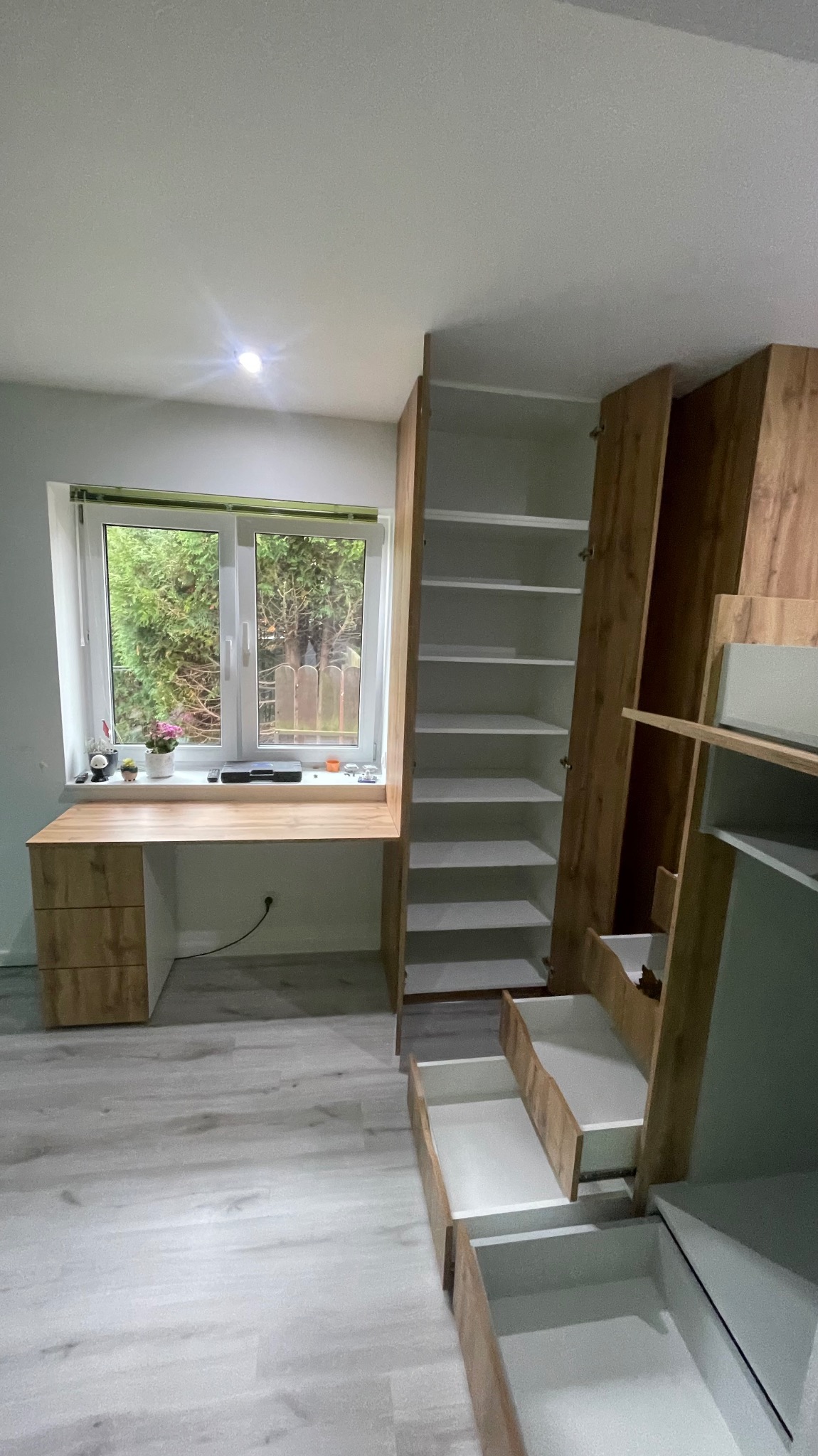 Nowoczesny pokój z biurkiem zintegrowanym z szafką pod oknem i wysoką szafą z otwartymi półkami oraz wysuwanymi szufladami, wykończony w odcieniach drewna i bieli.