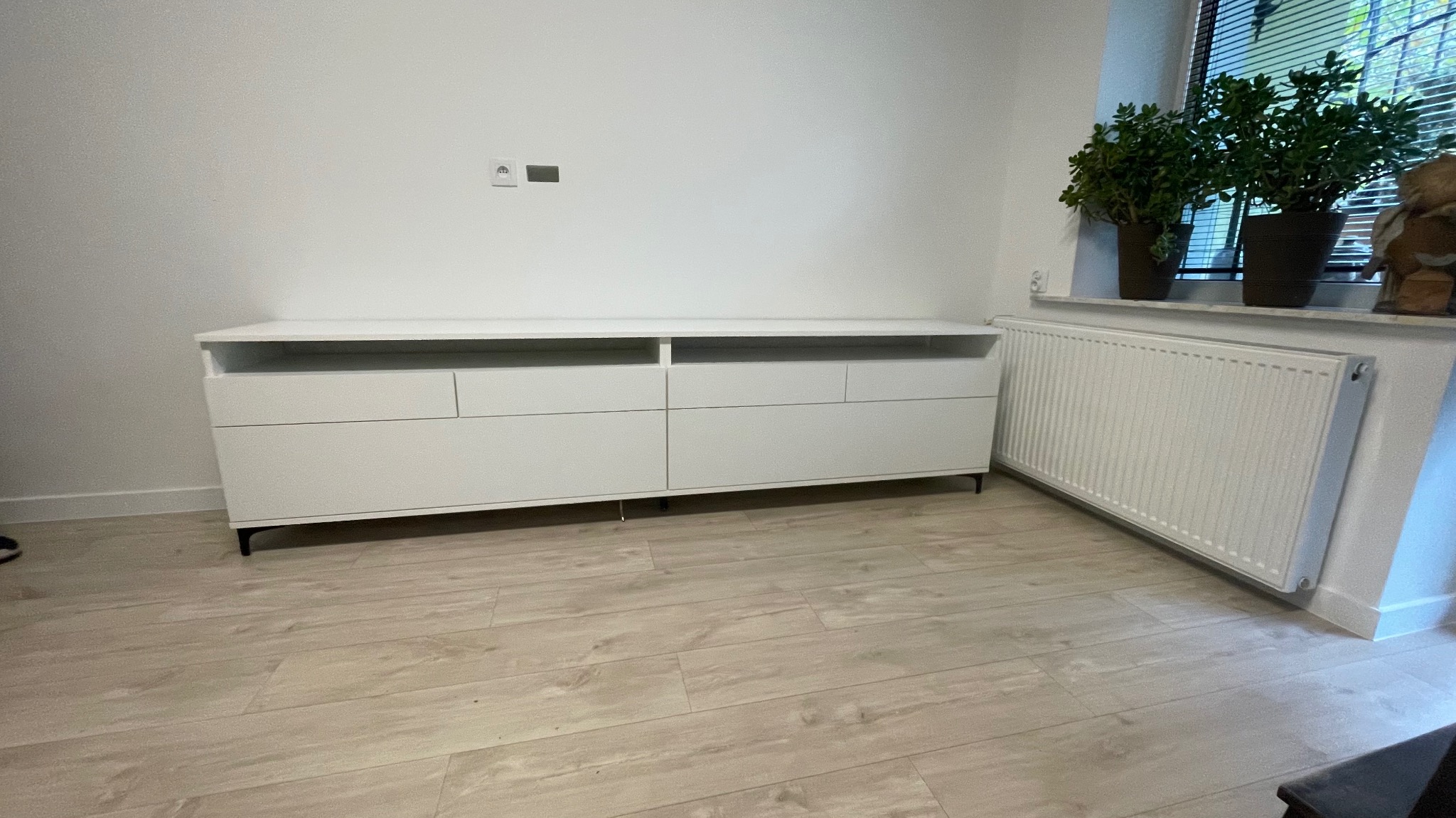 Biała komoda z szufladami i otwartymi półkami, ustawiona na drewnopodobnej podłodze, z grzejnikiem i roślinami w tle przy oknie z żaluzjami.