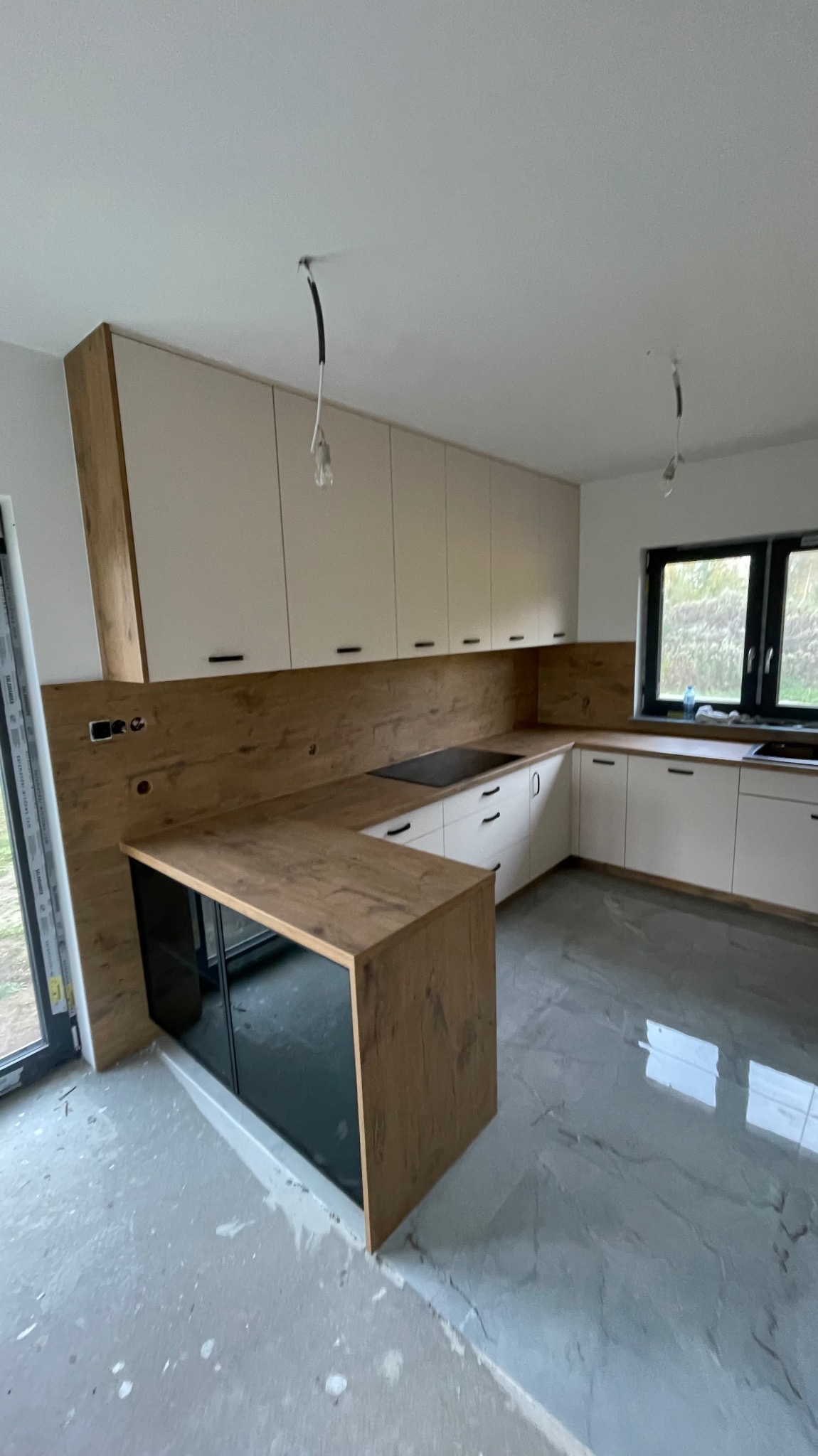 Nowoczesna kuchnia z białymi szafkami i drewnianym blatem, widoczne kable wystające z sufitu, sugerujące niedokończony montaż oświetlenia, podłoga w trakcie wykończenia.