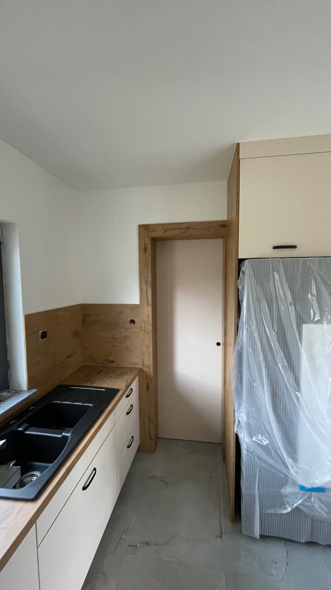 Nowoczesna kuchnia z białymi szafkami, czarnym zlewozmywakiem i blatem imitującym drewno, z widocznym fragmentem lodówki zabezpieczonej folią ochronną i drzwiami w drewnianej ościeżnicy.