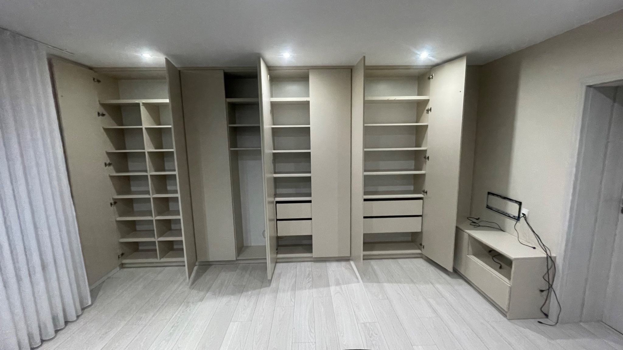 Jasna zabudowa meblowa z otwartymi szafami i szufladami, widoczny fragment zasłony i szafka RTV z wiszącym uchwytem na telewizor, biała podłoga.