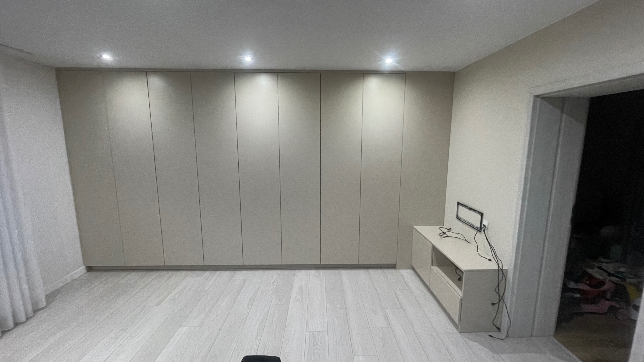 Beżowa szafa wnękowa z przesuwnymi drzwiami, zajmująca całą ścianę, oświetlona punktowymi lampami sufitowymi, na podłodze jasne panele, obok szafki RTV z czarną ramką na ścianie i widocznymi kablami.