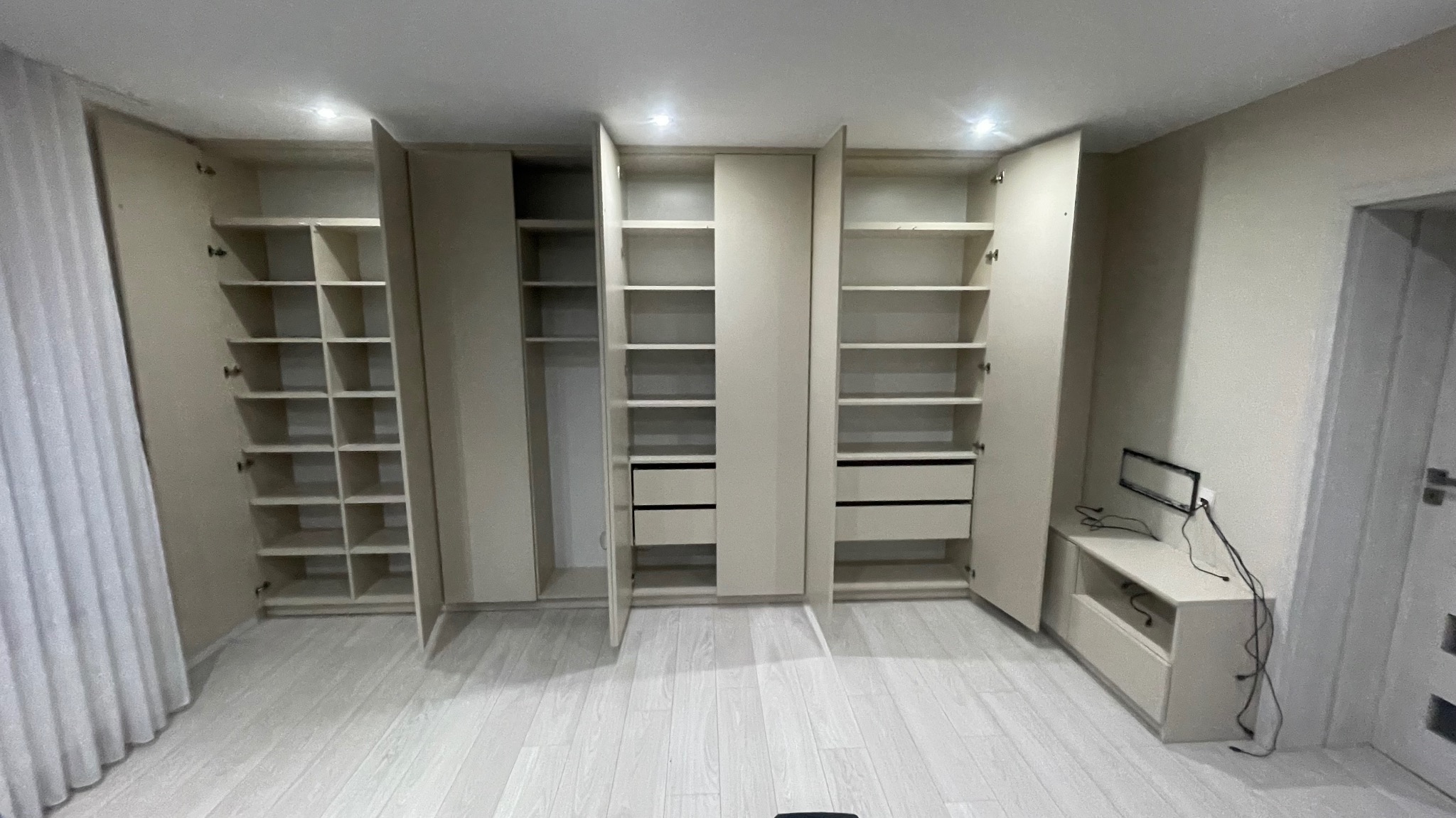 Jasna, nowoczesna garderoba z otwartymi półkami i szufladami, z widocznymi zawiasami i szczelinami między drzwiami, na tle jasnej podłogi i ściany.