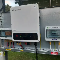 Biały falownik GoodWe zamontowany na metalowej konstrukcji z rozdzielnicami PV-DC i PV-AC, widoczne okablowanie i etykiety ostrzegawcze, tło z zielenią.