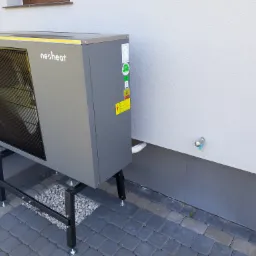 Szara pompa ciepła marki Nesheat, zamontowana na czarnej, metalowej konstrukcji, stojąca na szarej kostce brukowej obok ściany budynku z widocznym zaworem.
