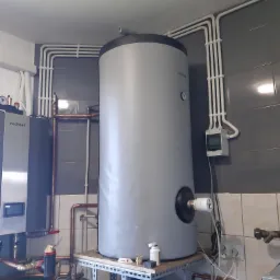 Szary, pionowy zbiornik na wodę z podłączonymi rurami miedzianymi i zaworem termostatycznym, zamontowany w kotłowni z szarymi kafelkami i białymi rurami elektrycznymi na ścianie.