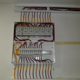 Rozdzielnica elektryczna z zabezpieczeniami przepięciowymi, okablowaniem oznaczonym żółtymi etykietami i listwami montażowymi w białej obudowie, zamontowana na białej ścianie.