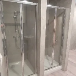 Dwie kabiny prysznicowe z przesuwnymi drzwiami w łazience wykończonej płytkami imitującymi beton, z widoczną armaturą i półkami wewnątrz.