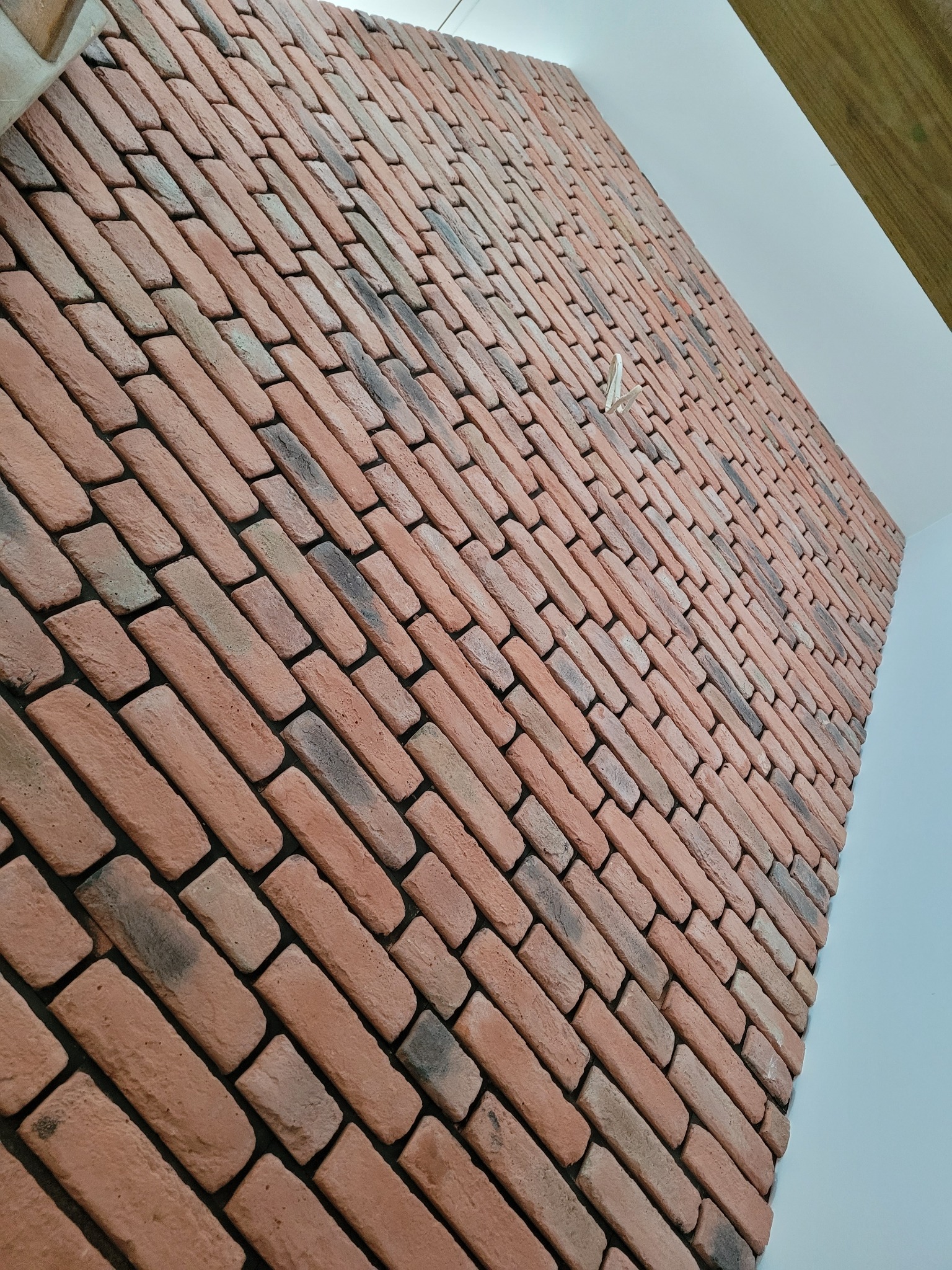 Ściana wykończona dekoracyjną cegłą klinkierową w odcieniach czerwieni i szarości, ułożona ukośnie, z widocznym fragmentem białego sufitu i drewnianej belki.