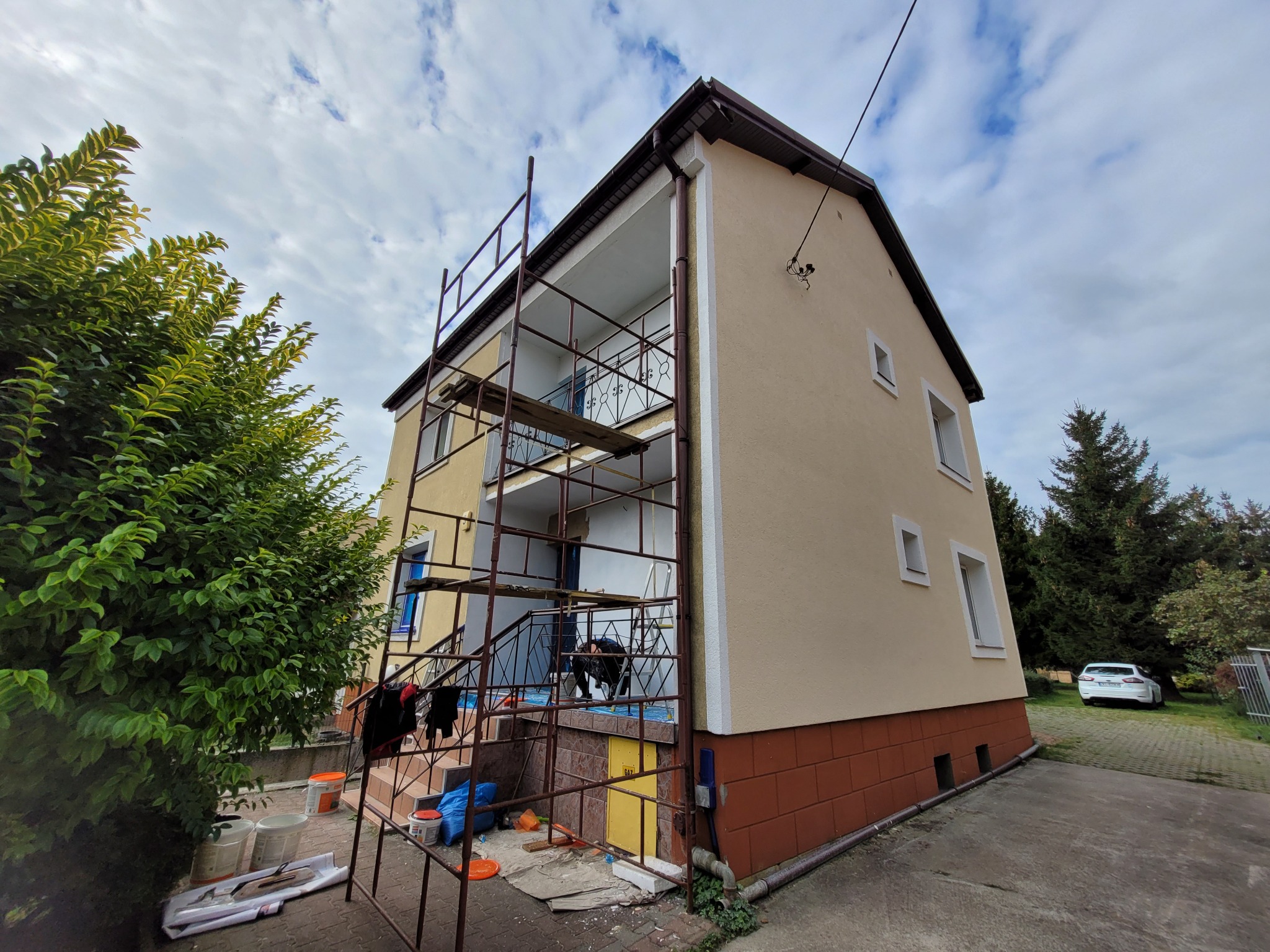 Dwupiętrowy dom z beżową elewacją i brązową cegłą na fundamencie, z metalowym rusztowaniem przy balkonie, widoczne narzędzia malarskie i wiadro z farbą, samochód w tle.