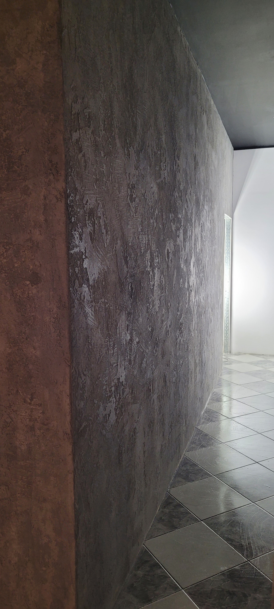 Dekoracyjna ściana z efektem betonu architektonicznego w odcieniach szarości, kontrastująca z fragmentem ściany w kolorze ceglastej czerwieni i szachownicą na podłodze.