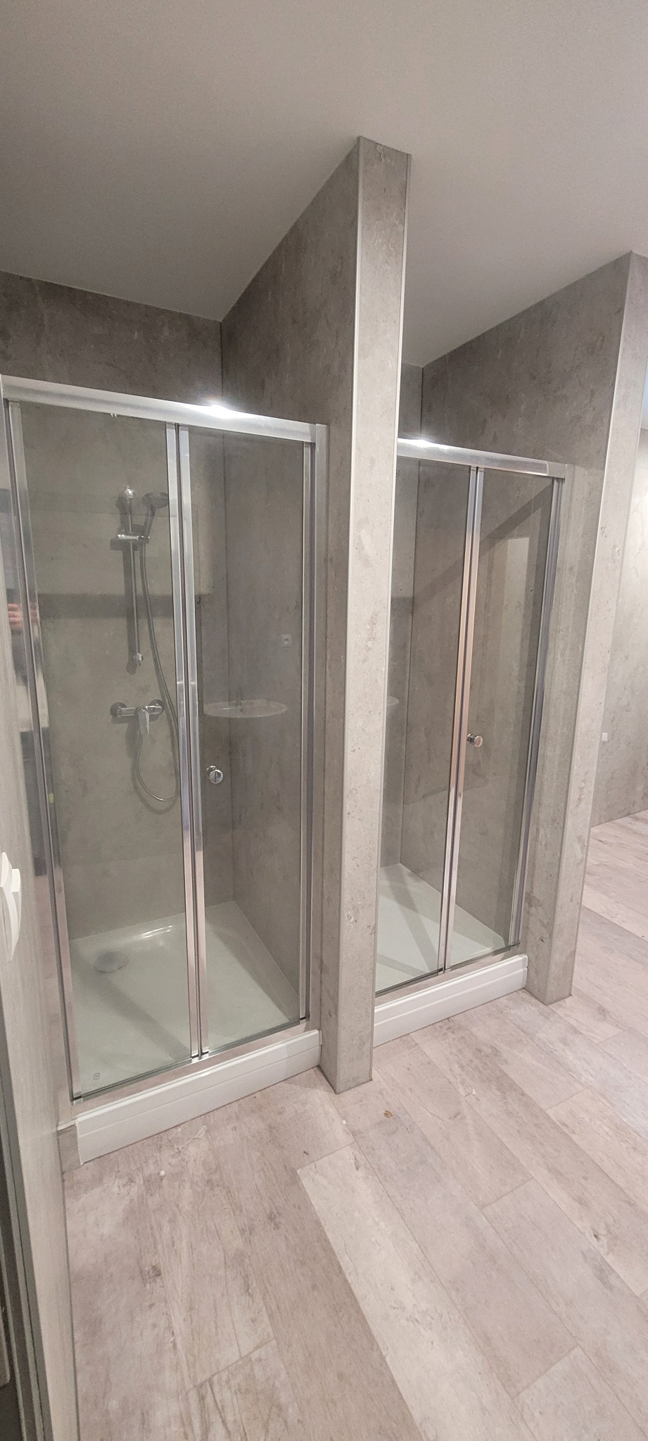 Dwie kabiny prysznicowe z przesuwnymi drzwiami w łazience wykończonej płytkami imitującymi beton, z widoczną armaturą i półkami wewnątrz.