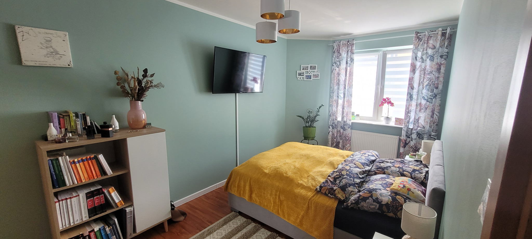 Jasna sypialnia z łóżkiem z żółtą narzutą, telewizorem na ścianie, regałem z książkami, zasłonami w kwiaty i trzema lampami wiszącymi.