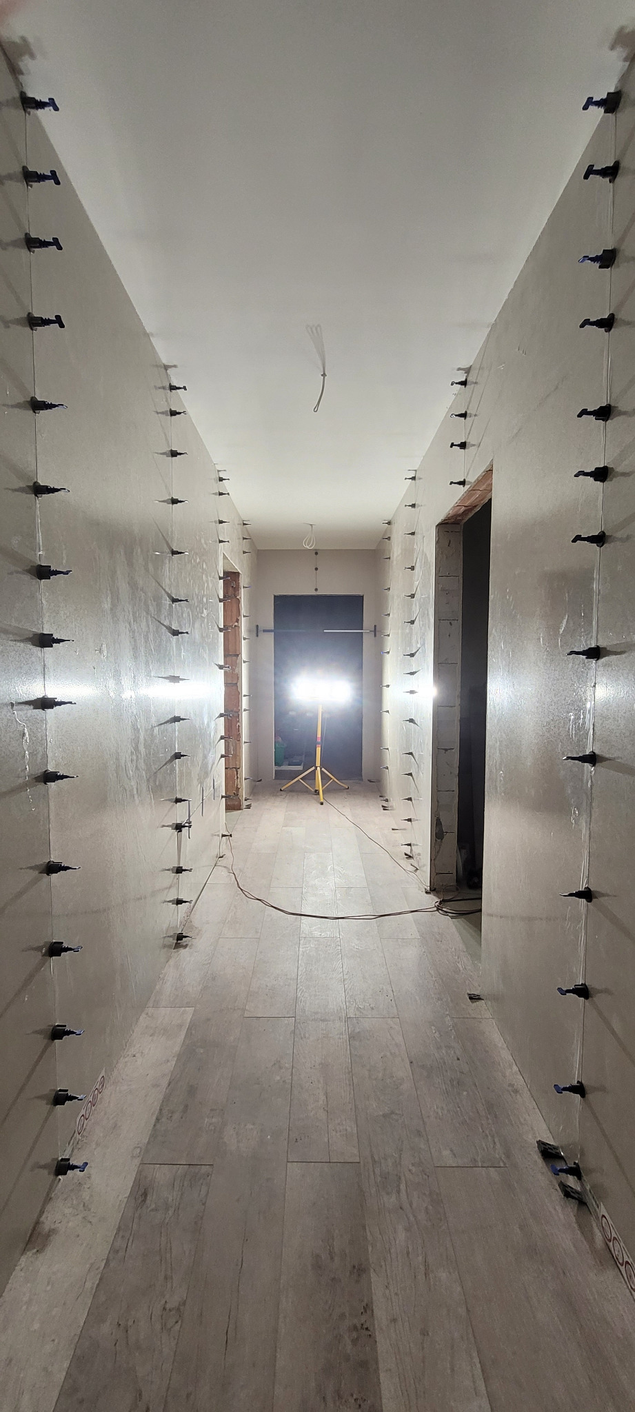 Korytarz w trakcie prac wykończeniowych, ściany przygotowane do malowania z użyciem systemu poziomowania płytek, widoczna lampa warsztatowa i płytki podłogowe imitujące drewno.