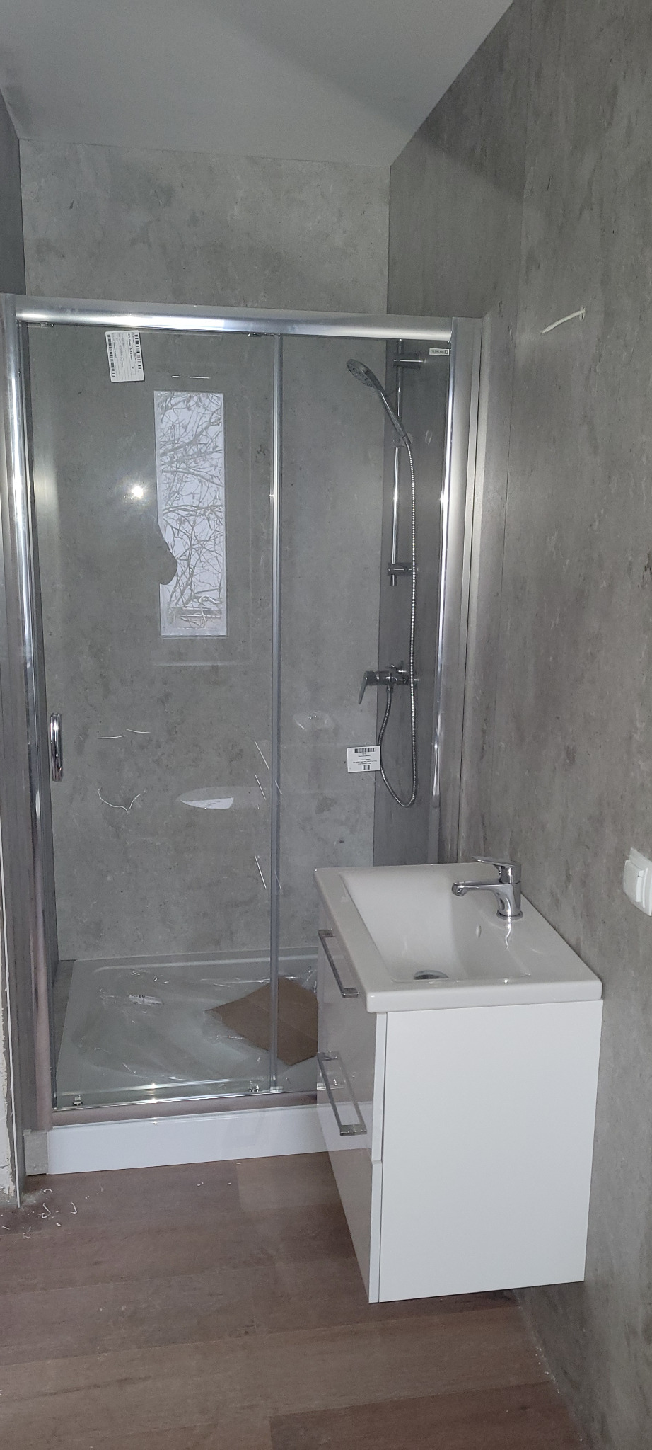 Nowa kabina prysznicowa z przesuwnymi drzwiami i biała szafka z umywalką zamontowane w surowym wnętrzu łazienki z szarymi ścianami.