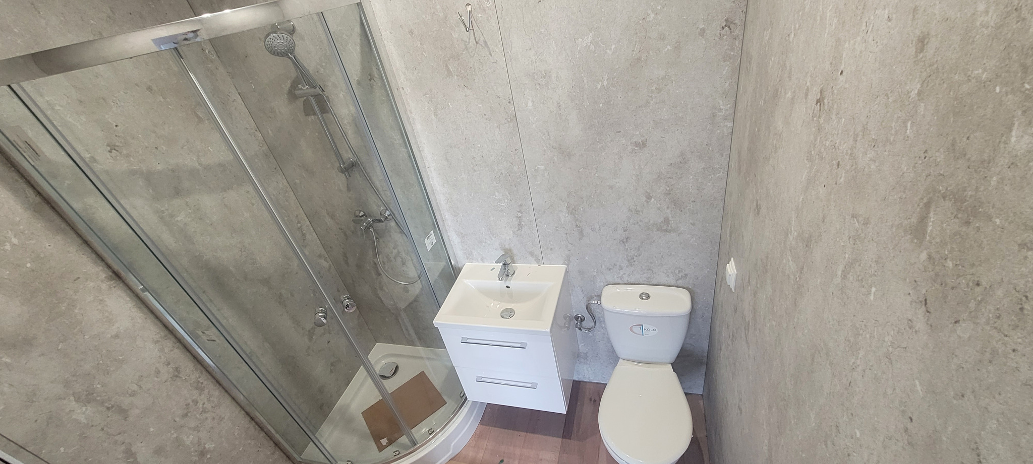 Nowoczesna łazienka z kabiną prysznicową, białą szafką z umywalką i toaletą w odcieniach szarości, wykończona płytami imitującymi kamień.