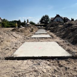 ALEXBUD ALIAKSEI MATSKEVICH - Przygotowane betonowe płyty z czerwonymi oznaczeniami ułożone w wykopie pod budowę, widoczny pracownik w oddali i błękitne niebo.