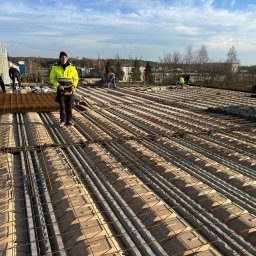 ALEXBUD ALIAKSEI MATSKEVICH - Prace zbrojarskie na budowie: widok z góry na ułożone zbrojenie na stropie żelbetowym z prefabrykatów, pracownicy w odblaskowych kamizelkach w tle, słoneczny dzień.