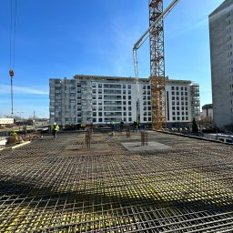ALEXBUD ALIAKSEI MATSKEVICH - Zbrojenie płyty fundamentowej budynku wielorodzinnego z widoczną konstrukcją stalową, żółtą izolacją i pracownikami w odblaskowych kamizelkach na tle miejskiego krajobrazu z dźwigiem i budynkami.