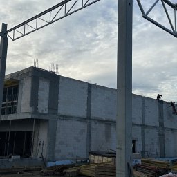 ALEXBUD ALIAKSEI MATSKEVICH - Wznoszenie dwukondygnacyjnego budynku z betonu komórkowego, widoczni pracownicy na podnośniku montujący szalunki, konstrukcja stalowa dachu w tle.